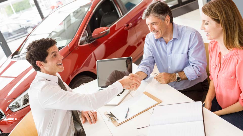 Ein Autoverkäufer schüttelt den Kunden die Hand in einem Autohaus in Lüdenscheid, im Hintergrund ist ein rotes Auto zu sehen und auf dem Tisch liegen Unterlagen.
